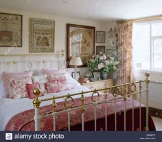 عکس - نمونه های عتیقه روی دیوار بالای تخت برنجی عتیقه با لحاف صورتی و کوسن ها در اتاق خواب کلبه ای با پارچه Toile de Jouy